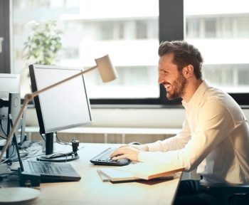 A man smiling while working at an office desk with a computer and natural daylight streaming in through large windows.
