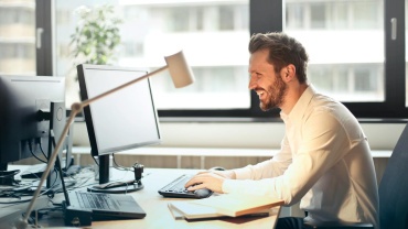 A man smiling while working at an office desk with a computer and natural daylight streaming in through large windows.